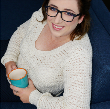 Woman in glasses holding a teal cup, sitting on a blue sofa, smiling at the camera. She's wearing a white sweater.